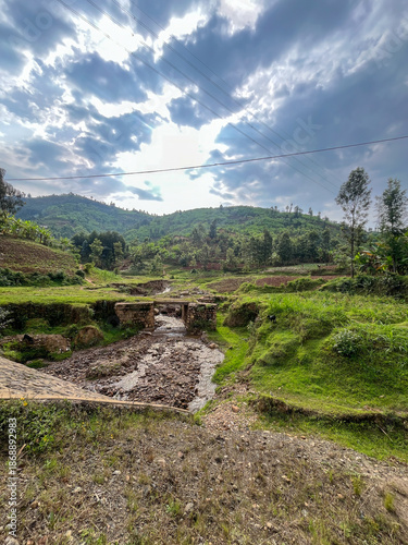 Wide view of a small river crossing with a stone bridge in a fertile green landscape under a dramatic sky in Rwanda.