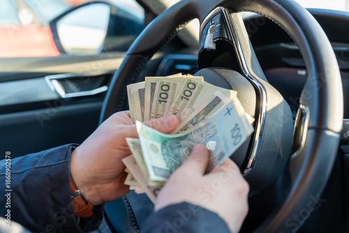 Man counting Polish zloty banknotes sitting in car behind steering wheel with hands holding cash money. Concept of car purchase, taxi driver income, fuel expenses and automotive business transactions.