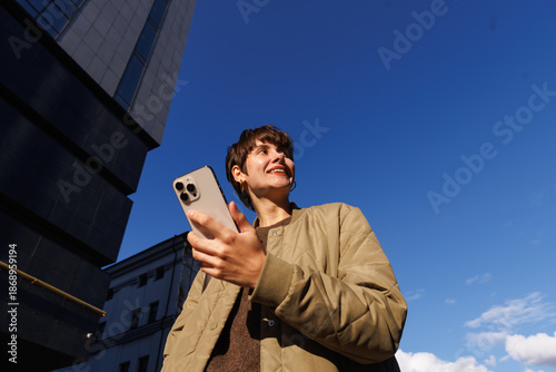 Smiling woman holding smartphone in city sunlight against blue sky