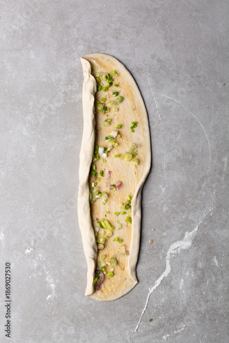 Overhead view of raw chinese scallion pancakes being rolled on grey countertop, top view of homemade scallion pancakes being shaped on countertop, process of making cong you bing