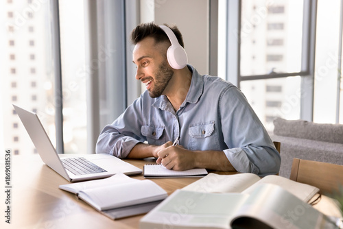 Tableau sur toile Young 30s Hispanic man listen to audio class using modern wireless headphones, taking notes in notebook, engaged in online course, participate in webinar for gaining new knowledge remotely