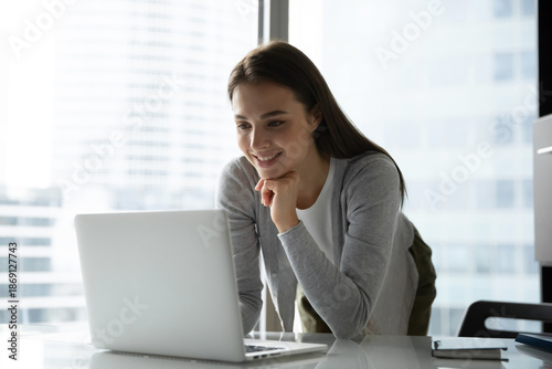 Inspired specialist at work. Smiling young female business professional executive stand leaning at office desk use notebook for videocall communication interact via text chat review online content