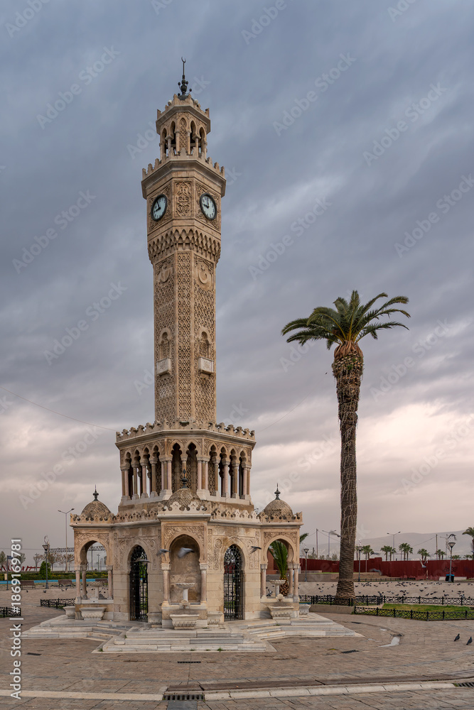 Fototapeta premium Iconic Izmir Clock Tower, a historic landmark, stands tall with a palm tree in Konak Square, Izmir, Turkey, under a dramatic cloudy sky. Pigeons gather on the ground.