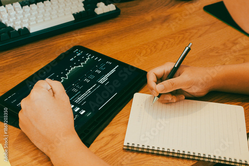Investor analyzing stock market charts on digital tablet while taking notes in notebook, financial trading and data analysis concept.