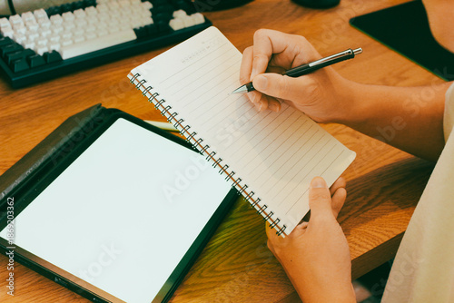 Man holding spiral notebook and pen over blank white screen tablet on wooden desk, mockup and creative workspace concept.