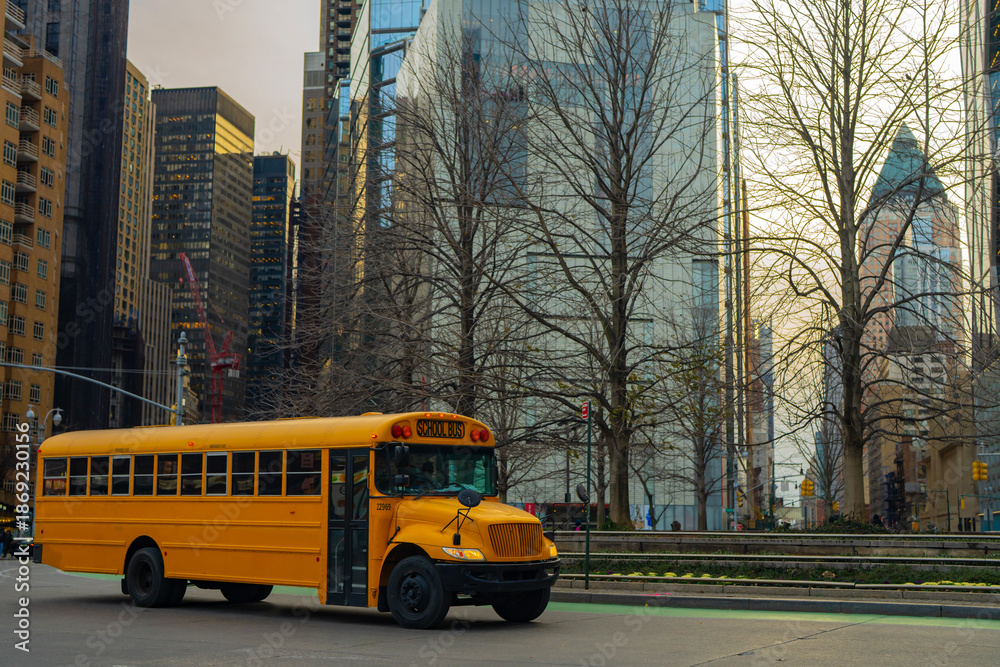 Obraz premium NYC school bus driving through Manhattan street. New York City yellow school bus in urban traffic. NYC transport classic yellow school bus. Manhattan city street with NYC school bus.