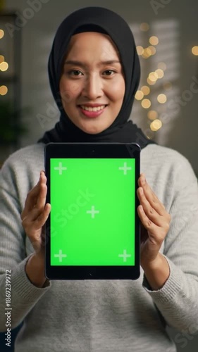 A smiling woman wearing a hijab holds up a tablet with a green screen in a cozy indoor setting.