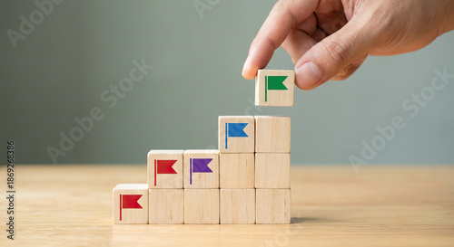 Human hand placing green flag block on top of ascending wooden steps, symbolizing business success.