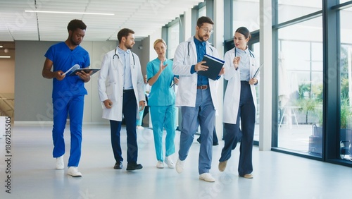 Medical staff walking calmly along bright corridor inside modern hospital. Focused doctors reviewing notes while discussing plans. Nurses joining conversation and moving together during routine moment