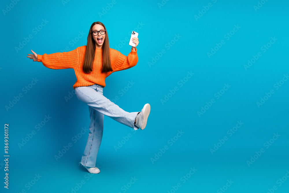 Fototapeta premium Young woman in orange sweater jumps with a smile holding phone against bright blue background fashion lifestyle image