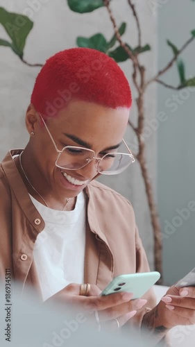 Red hair African American woman smiling uses smartphone and credit card for online shopping indoors with houseplant background, checking messages relaxed modern lifestyle