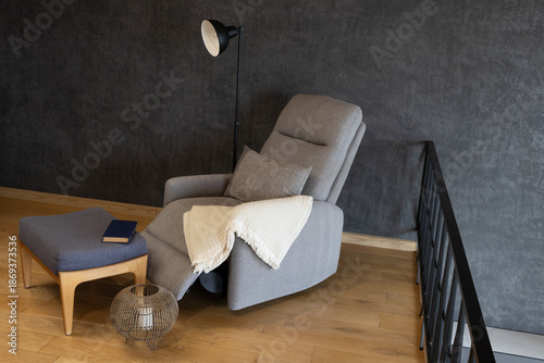 Tableau sur toile Modern cozy interior featuring a grey recliner armchair with a white knit throw blanket and a decorative pillow, accompanied by a grey footstool, and a black floor lamp against a dark textured wall
