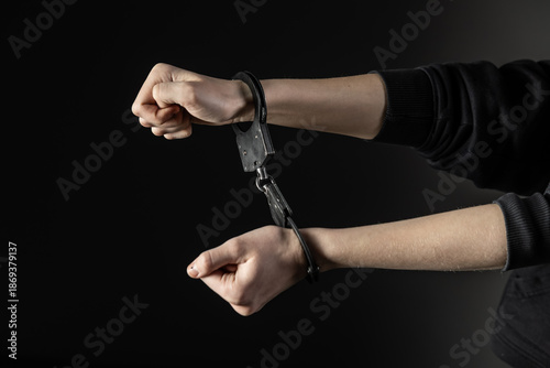 Fotografie A close-up of a person’s wrists locked in black handcuffs, set against a dark background