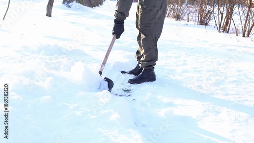A person shoveling snow in a winter landscape. The individual wears dark pants and boots, working to clear a path in the white snow.