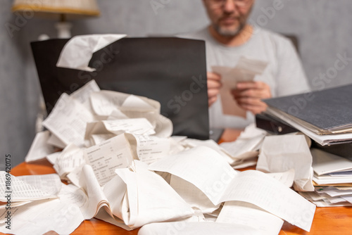 Overwhelmed man reviewing receipts behind a desk covered with scattered papers and folders. Concept of financial stress, expense tracking, paperwork overload, and budgeting problems.