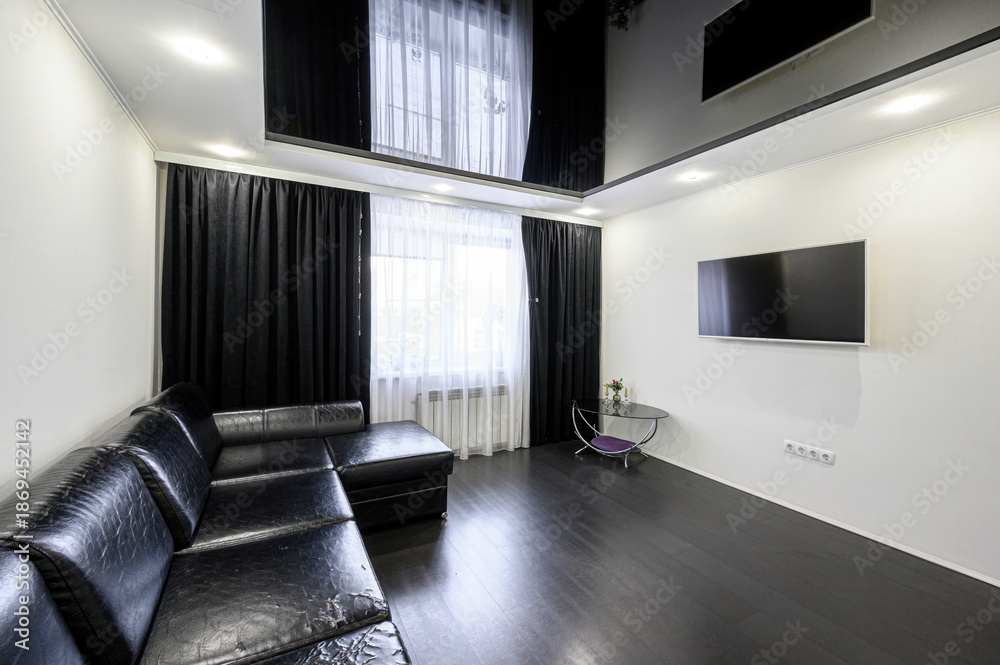 Fototapeta premium Modern living room with black leather sectional sofa, wall-mounted TV, dark wooden floor, black curtains, sheer white drapes, small glass side table with decor
