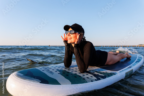 Smiling woman enjoys surfing on Baltic Sea waves, laughing joyfully lying on board with splashing water around. Bright sunlight and her laughter reflect pure joy, freedom and vibrant summer adventure
