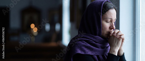 Devout woman wearing purple headscarf praying with clasped hands in church. Christian concept of Lent, faith and repentance. Spiritual portrait near window with icons in background.