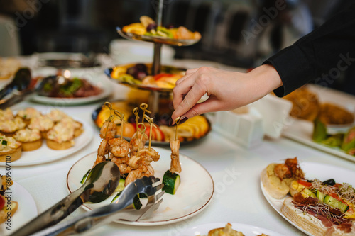 A photograph of a woman holding a canapй of meat against a round table with a variety of fresh food at a banquet or buffet. Catering concept, buffet.