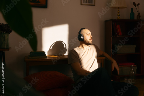 A man relaxes on the floor of a sunlit room while wearing headphones with his eyes closed. The image captures a quiet moment of digital connection and leisure in a cozy, modern home environment.