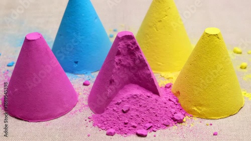 Group of five conical sand sculptures in vibrant pink blue and yellow hues arranged on a textured fabric surface with loose colorful sand scattered around them in a studio setting with soft lighting