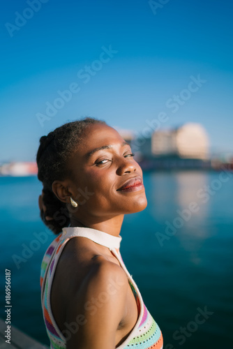 Canvas Print Confident young woman smiling softly toward camera, radiating calm energy and empowerment while enjoying peaceful waterfront moment under blue sky