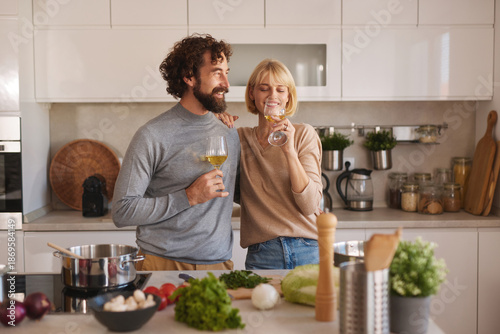 Canvas Print A couple stands in a stylish kitchen, smiling and holding glasses of wine as they prepare a meal