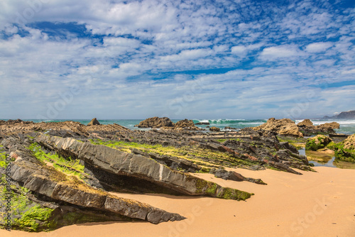 A panoramic view of Praia do Castelejo in the Algarve, Portugal, showcasing its wild, untamed beauty with golden sands meeting jagged, seaweed-draped rock formations under 