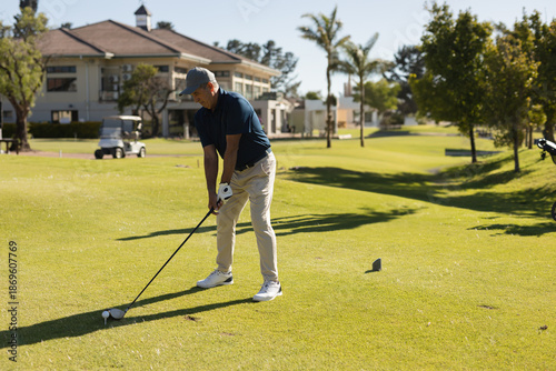 Senior man aligning driver on tee box near clubhouse with golf cart and ball, copy space