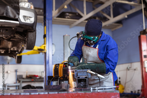 African American woman wearing goggles using grinder over vise sparking near car lift at workshop