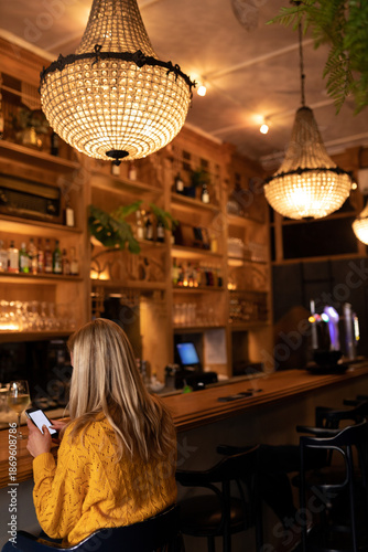 Woman wearing yellow sweater sitting at bar counter using smartphone near wine glass, copy space