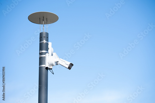 Modern CCTV surveillance camera mounted on a metal pole against a clear blue sky. Outdoor security system with copy space, minimalistic composition and clean background.