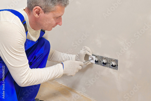 white-haired man in blue overalls is fixing a wall outlet