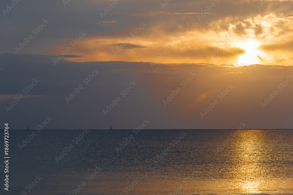 Naklejka premium Sailboat on the horizon under a gray and orange sky with the sun peeking through clouds