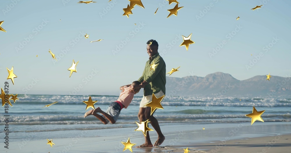Fototapeta premium Swinging father in green-top light-shorts holding son over beach waves distant range gold stars