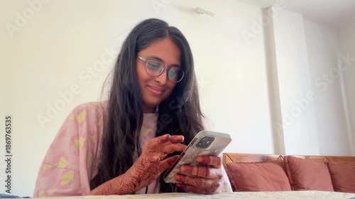 Young Indian woman in casual clothing sitting in a living room and scrolling on her smartphone, Modern lifestyle 