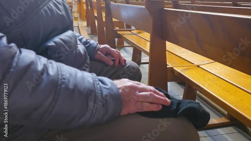 A man sits quietly on a wooden pew of church holding a black cap in his hands