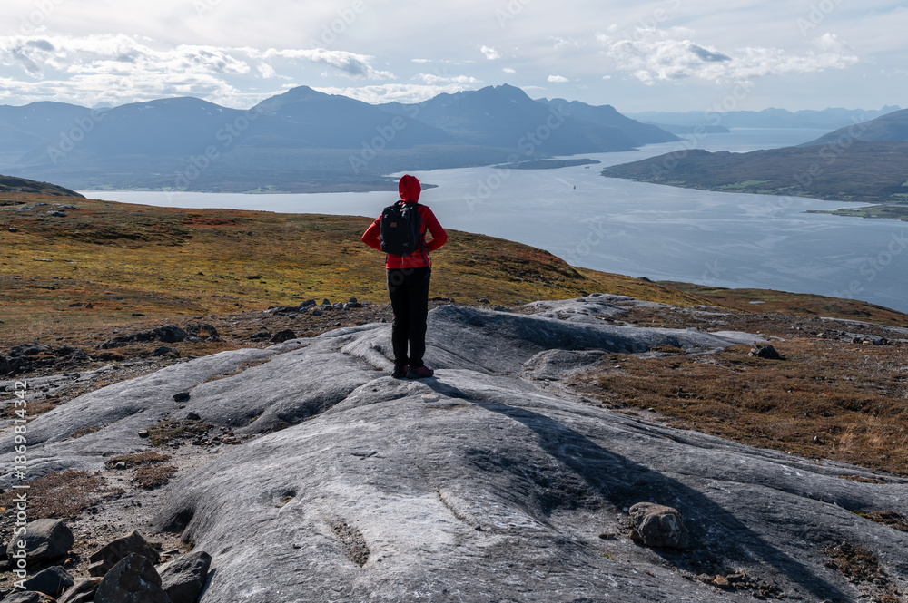 Fototapeta premium Young woman hiker standing on top of a mountain overlooking the Arctic city of Tromso and Norwegian fjords in autumn.