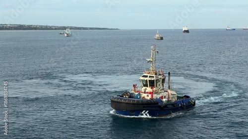 A tugboat works in a busy waterway close to the city harbor. It navigates the water as ships await assistance. Workers are seen on board, managing operations.