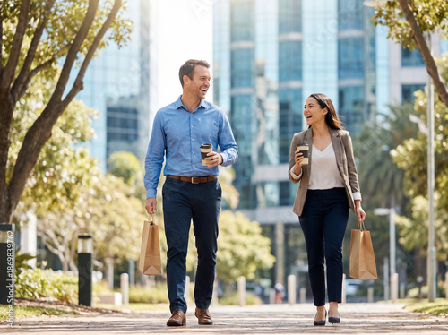 Smiling business colleagues walking outdoors with coffee and lunch bags, laughing together during a sunny city break near modern office buildings.