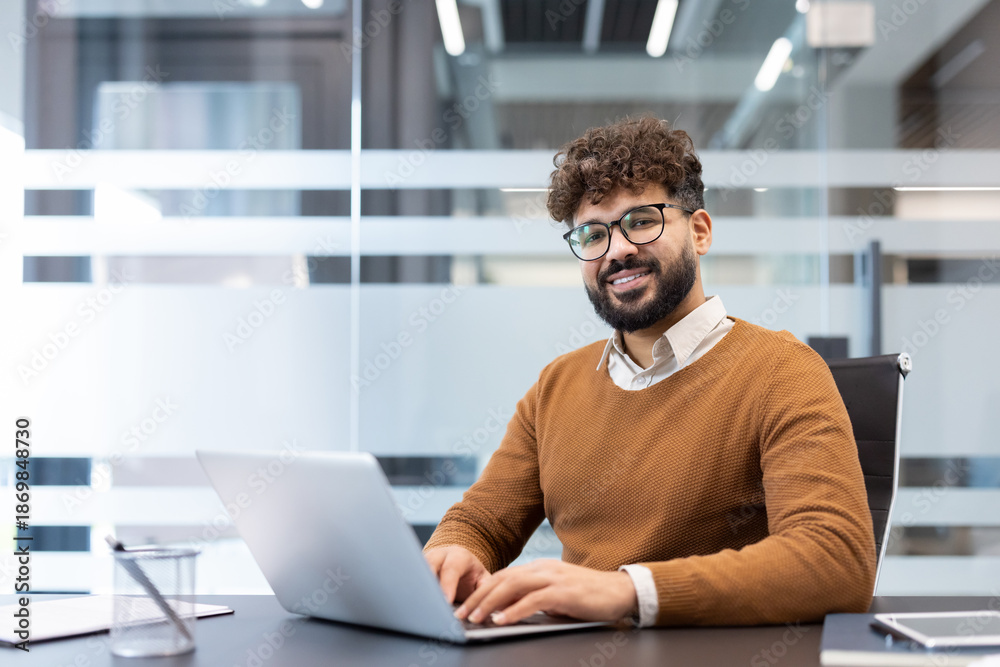 Obraz premium Young man with beard and glasses smiling at camera, sitting at a desk and using a laptop in a contemporary office environment, embodying professionalism and success