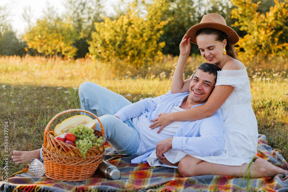 Fototapeta premium A couple relaxes on a blanket in a sunny field, sharing laughter and enjoying a picnic with a fruit basket, surrounded by nature