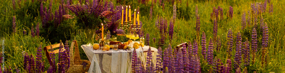 Obraz premium Banner. Beautiful romantic outdoor wedding decor. Table decorated with purple lupines flowers, lemons. Wineglasses with limoncello. Sunset, summer, golden hour. Perfect surprise date for loving couple