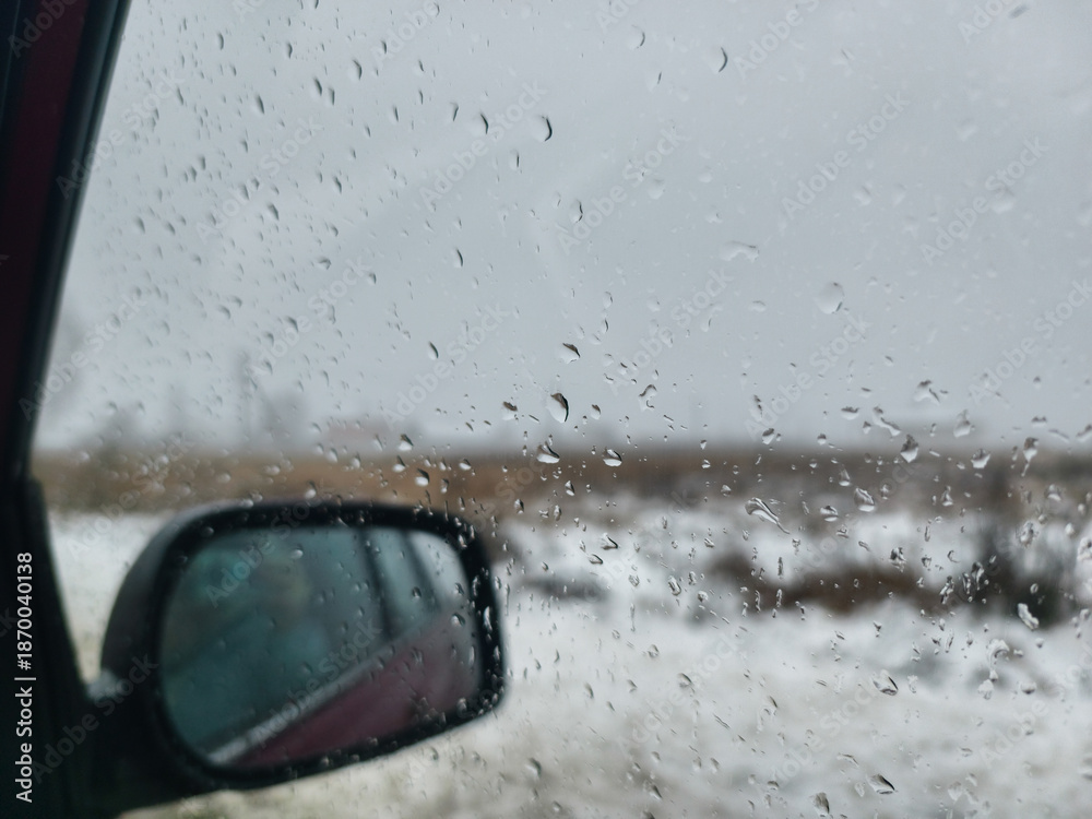 Fototapeta premium Raindrops collecting on the car window blur the view of the snowy winter landscape. The car's side mirror reflects the scene under the gray sky, creating a gloomy atmosphere.