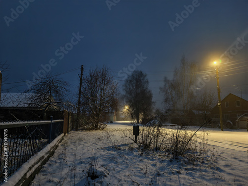 Night falling over a snow-covered rural street as warm lamplight from streetlights and houses glows against a dark blue winter sky, creating a peaceful, solitary scene.