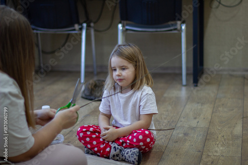 A mother and a small child sit on the floor, making a craft out of a stick with scissors. The family spends time being creative at home, encouraging the child's early development and imagination.
