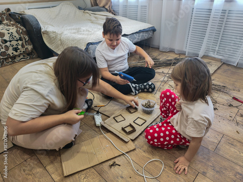 A mother and children are sitting on the floor, enjoying time together and crafting. They cut and glue natural materials, such as twigs and cardboard, for a creative activity.