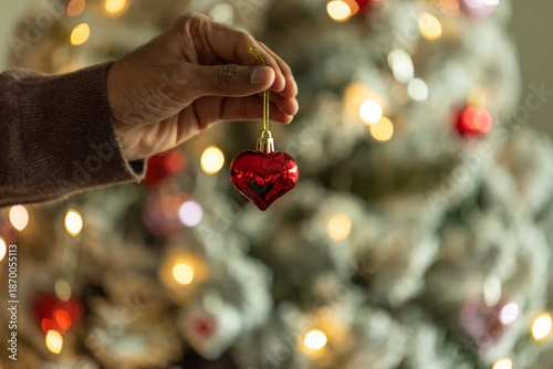 Hand holding red heart ornament by christmas tree