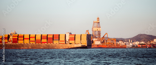 Cargo container ship passing an offshore oil rig near coastal port city at sunset, stacked containers on deck, calm sea and skyline, global trade and energy industry