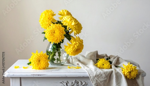 Bright yellow pompom chrysanthemums in a glass vase on a white table with a linen cloth